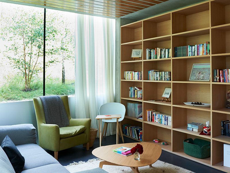 A warm reading room inside a Maggie's Centre: a green armchair, shelves of books, and a full-height window looking onto birch trees