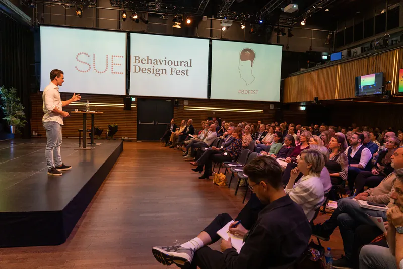 Spreker op het podium met de SUE Behavioural Design Fest-schermen en het publiek