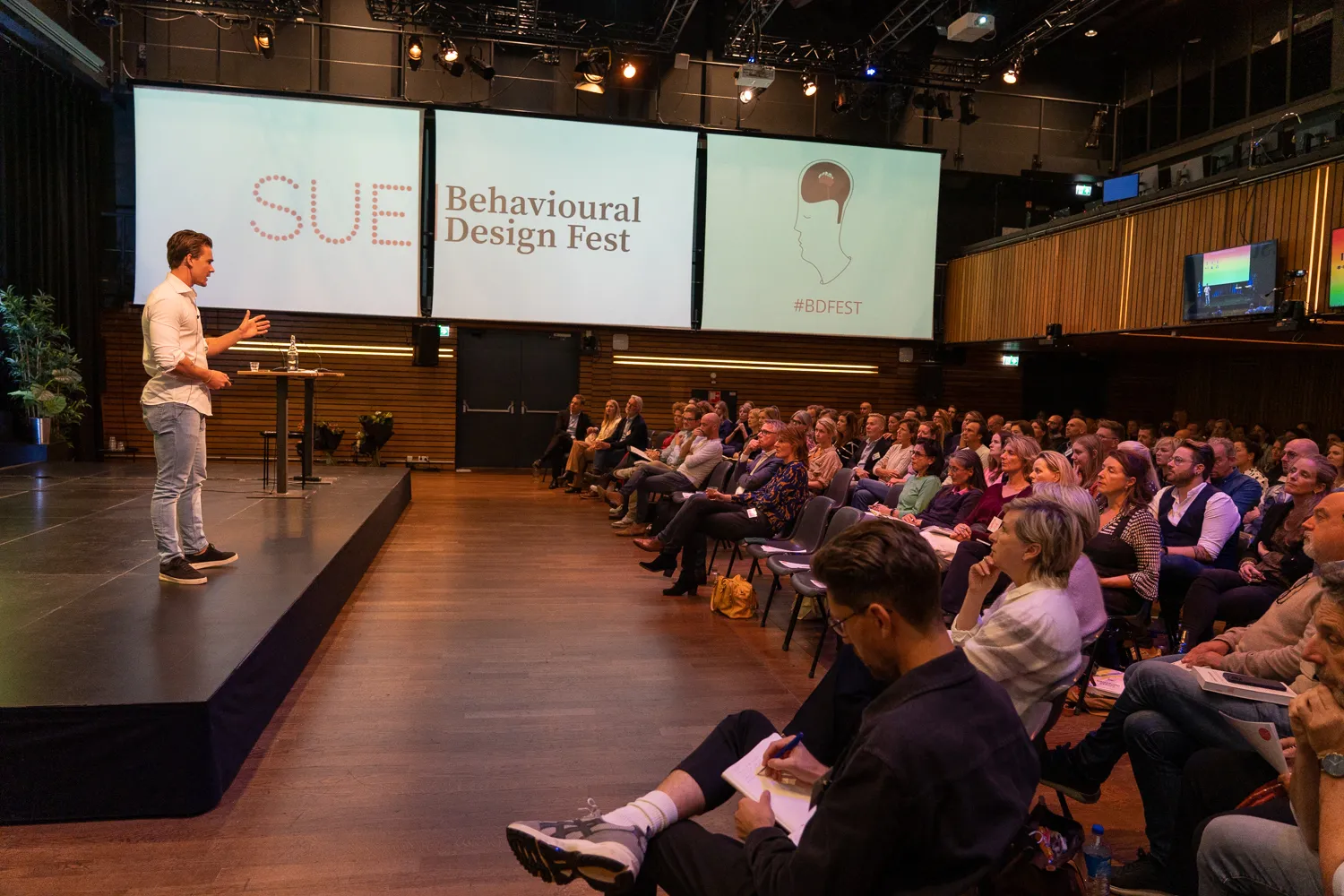 Spreker op het podium met de SUE Behavioural Design Fest-schermen en het publiek