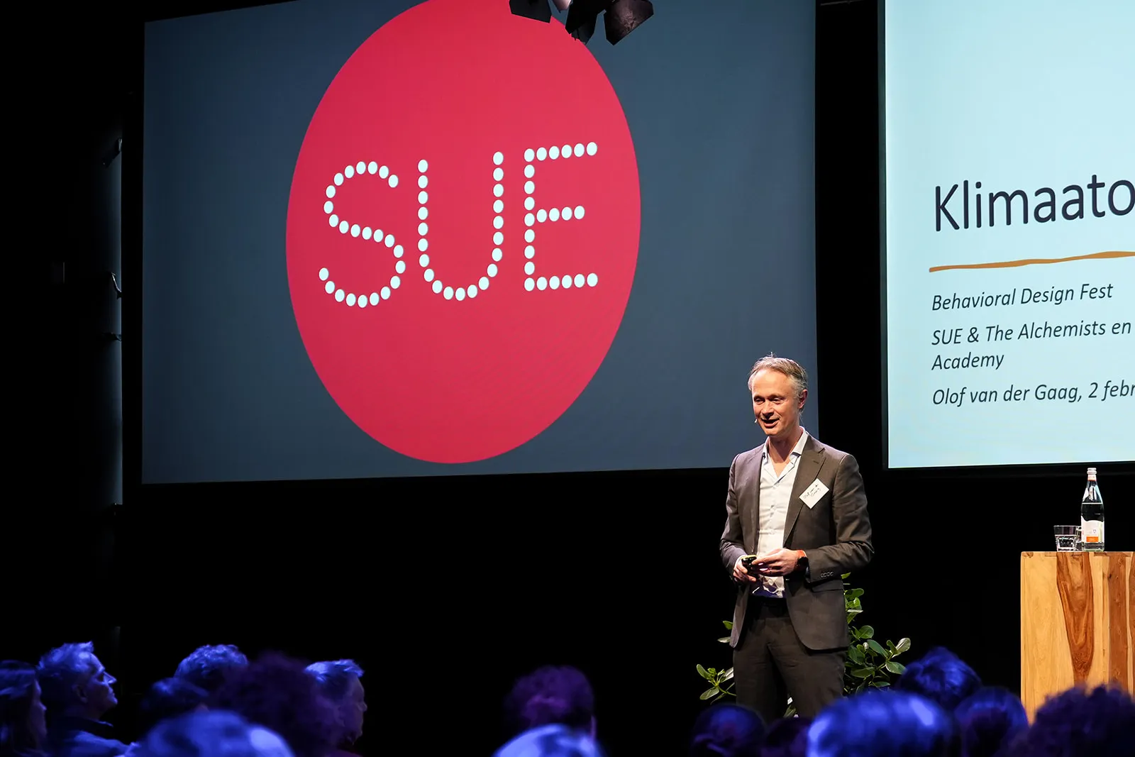 Spreker op het podium van het Behavioural Design Fest met het rode SUE-logo groot op het scherm