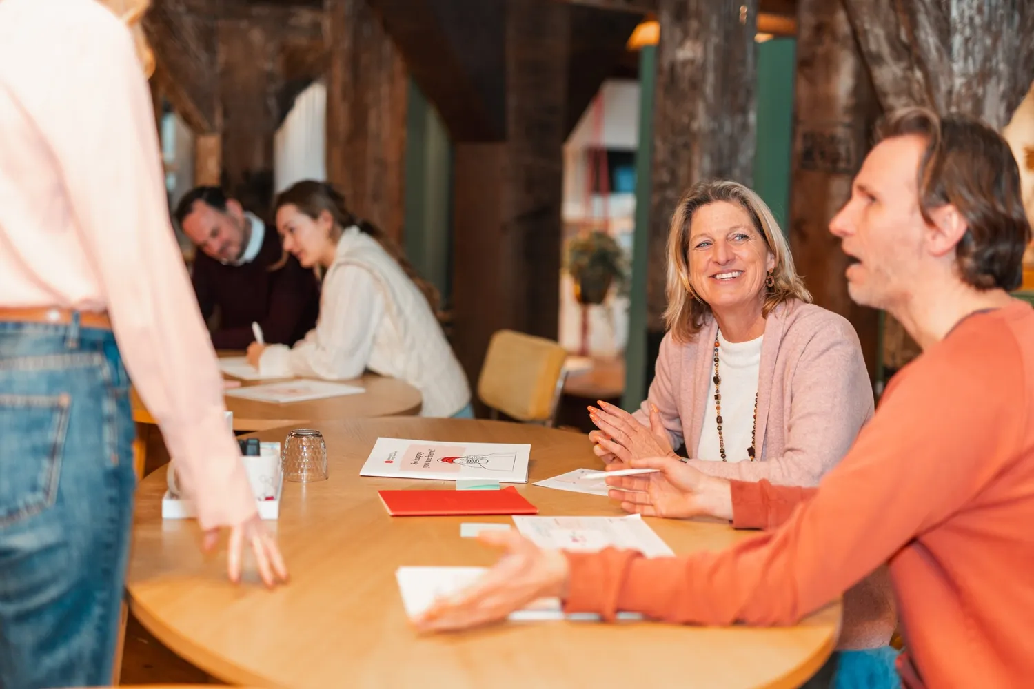 Participants in conversation at two work tables with SUE worksheets; in the background a colleague takes notes