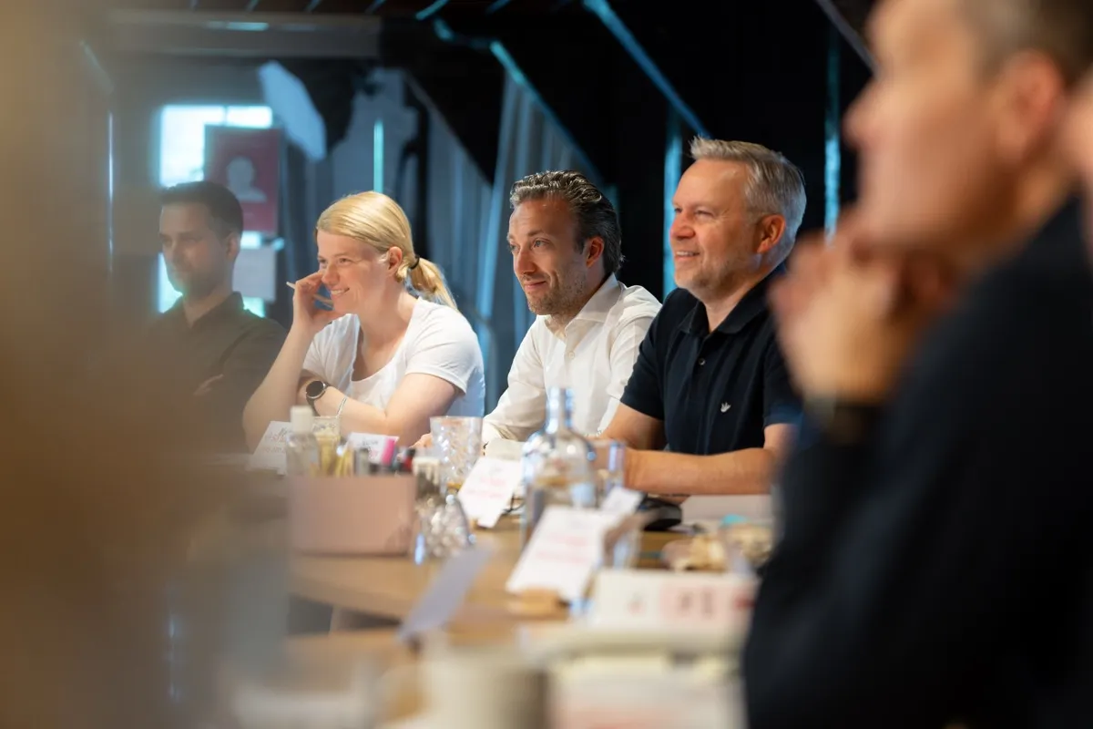 Een rij deelnemers aan de tafel in de SUE-zaal luistert naar de trainer, twee van hen glimlachen