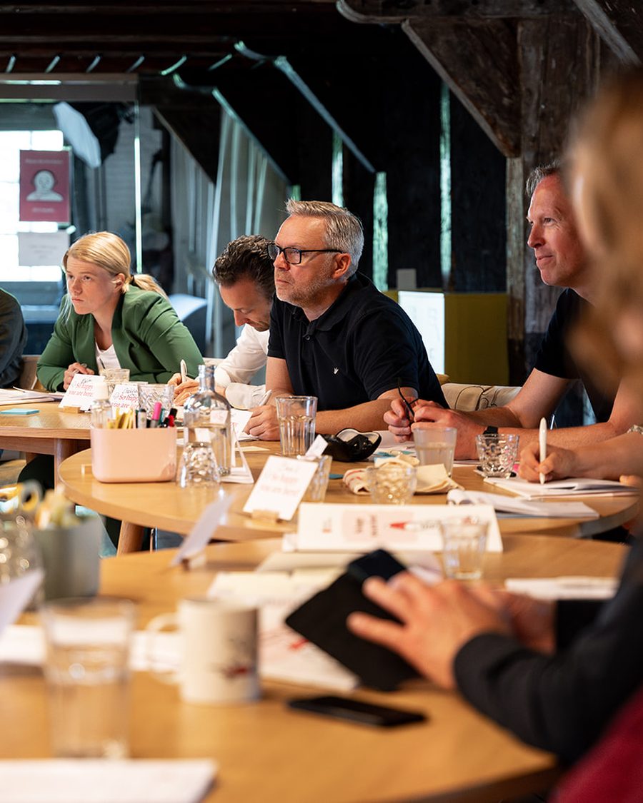Deelnemers aan een SUE-training werken samen aan een lange houten tafel in het Amsterdamse trainingslokaal, met werkbladen en aantekeningen voor zich.