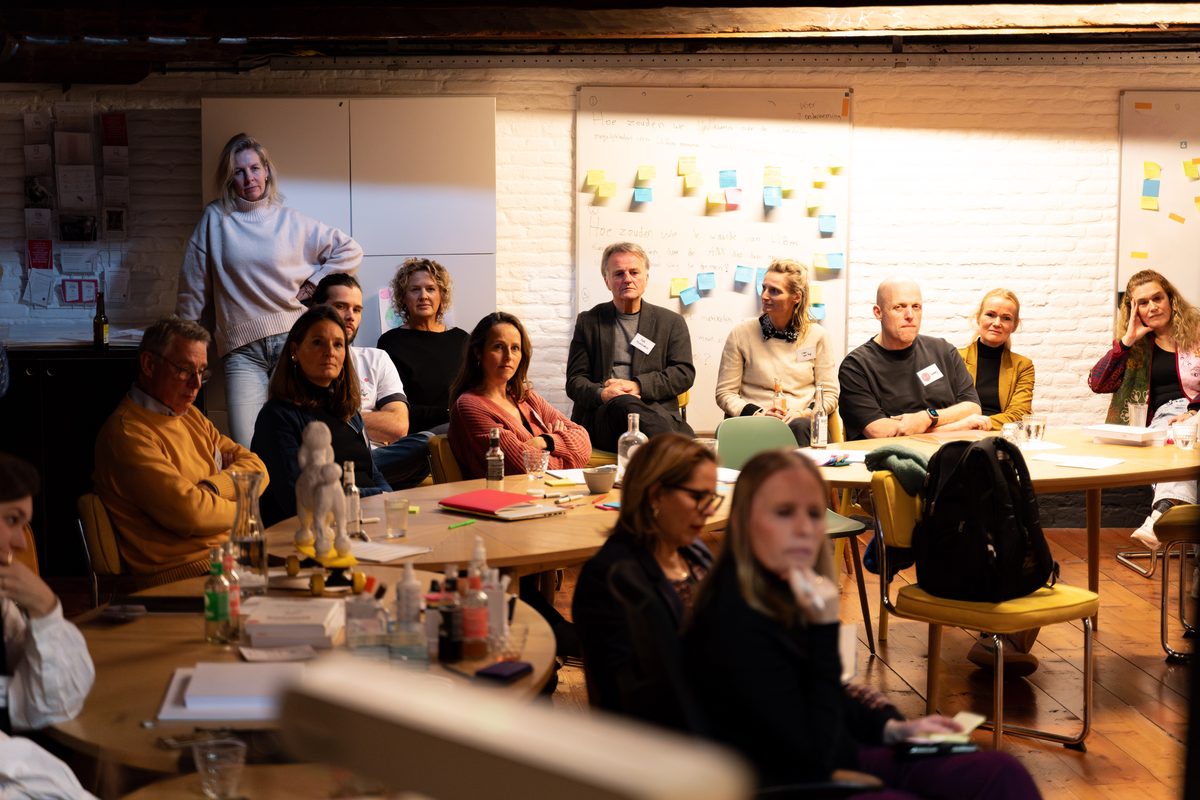 Een volle trainingsruimte bij SUE in Amsterdam: zestien deelnemers rond de tafels, met post-its aan de wand achter hen.